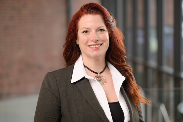 A woman with long red hair, wearing a dark blazer over a white collared shirt and a statement necklace, smiles at the camera. The blurred background hints at large windows and a brick wall, reflecting her professionalism as part of the DeCotis Team in real estate.