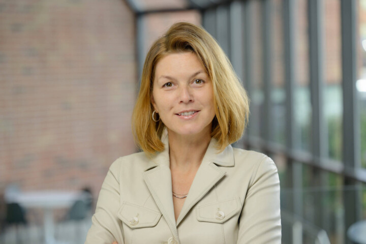 A woman with shoulder-length blonde hair, wearing a light beige blazer, stands indoors with her arms crossed and smiles at the camera. The background features large windows and a brick wall, reflecting the professional spirit of the DeCotis Team in real estate.