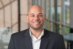 Frank Moran's headshot with a blurred out background of a brick wall and windows. He is wearing a dark sport coat and white collared shirt and he is smiling.